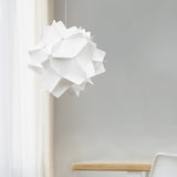 A white matte pendant lamp shade with a geometric pattern, hanging from a ceiling in a room with a wooden chair and a white curtain.