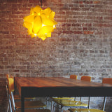 A yellow glossy pendant lamp shade with a geometric design, hanging over a wooden table in a room with brick walls and yellow chairs.